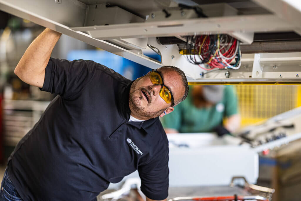 Worker in black polo shirt looking up at exposed wiring and electrical components during maintenance inspection of industrial conveyor equipment.