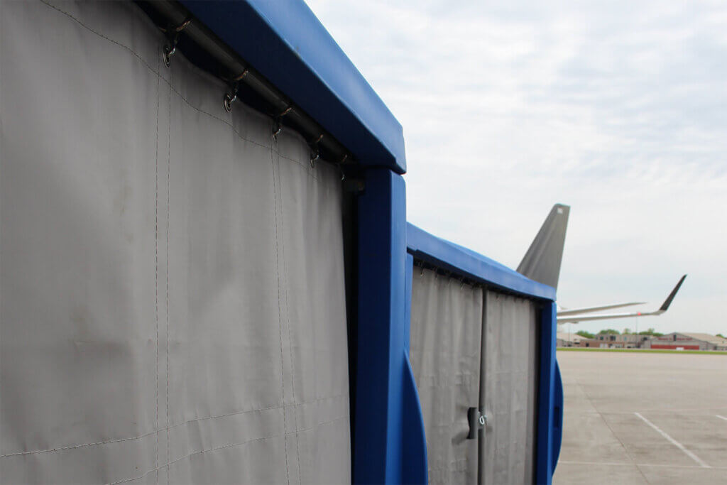Blue baggage cart with gray curtains on an airport tarmac with aircraft tail visible in background.