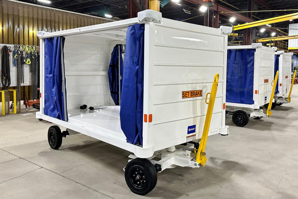 White airport baggage cart with blue curtains and yellow tow bar in a manufacturing warehouse.