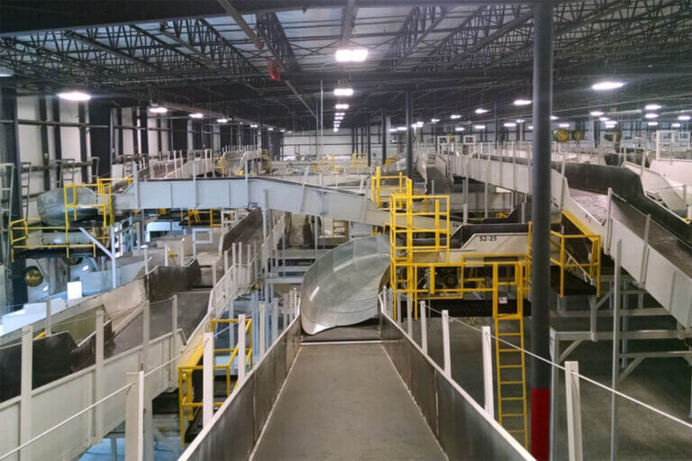 Complex network of conveyor belts, chutes, and platforms with yellow safety railings in a large warehouse facility.