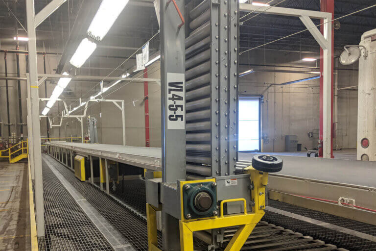 Industrial conveyor system with a yellow-framed transition roller in vertical storage position at loading dock station