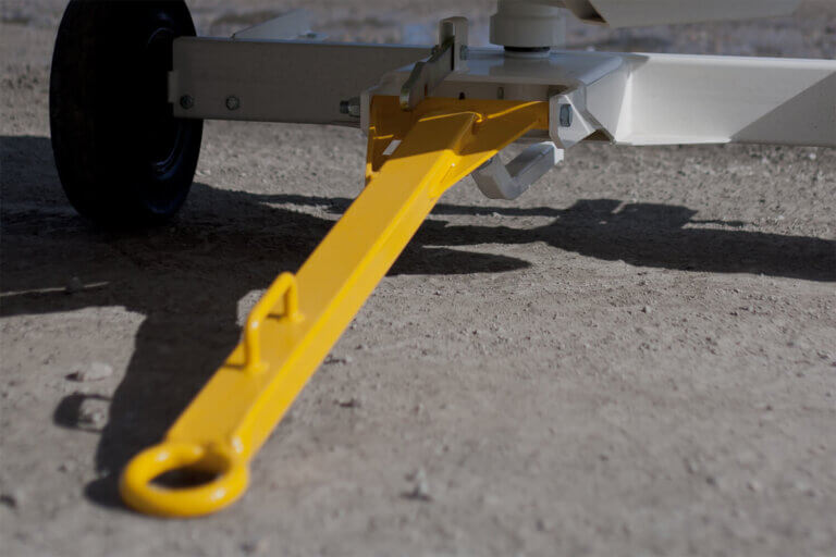 Yellow towbar extended in operational position attached to white baggage cart on gravel surface.
