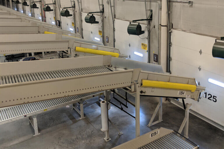 Series of gravity roller conveyors with yellow bumper guards installed at multiple loading dock doors.