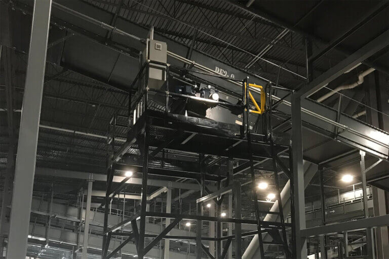 Elevated black metal platform supporting industrial control equipment in a large manufacturing facility with high ceilings.