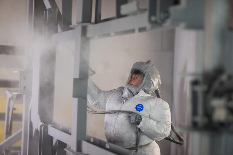 Factory painter wearing white Tyvek protective suit and respirator applying powder coating to metal framework.