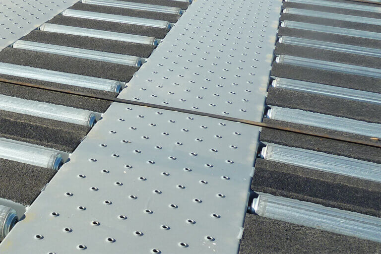 Metal walkway surface with perforated panels over cylindrical rollers for material movement.