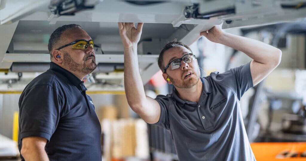 Two technicians in gray and navy polo shirts examining overhead equipment in manufacturing facility.