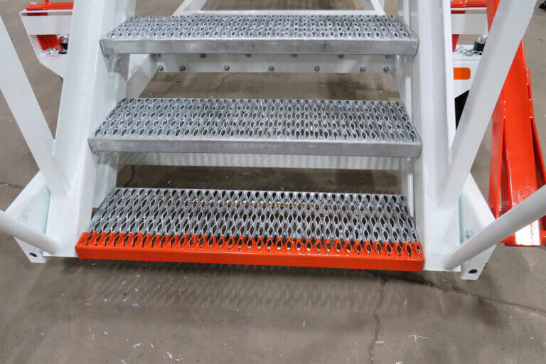 Three metal stair steps with diamond-pattern safety grating and orange safety edge on the bottom step.