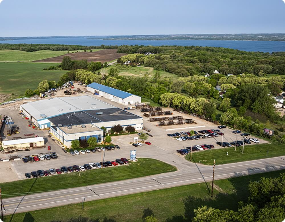 Bird's eye view of industrial facility with white buildings and parking lots surrounded by green fields and trees with a large lake visible in the background.