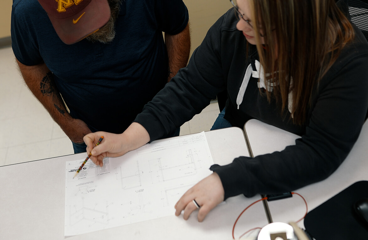 Two people in dark clothing reviewing engineering blueprints at a desk, with a person in a cap and another person with long hair marking the document with a pencil.