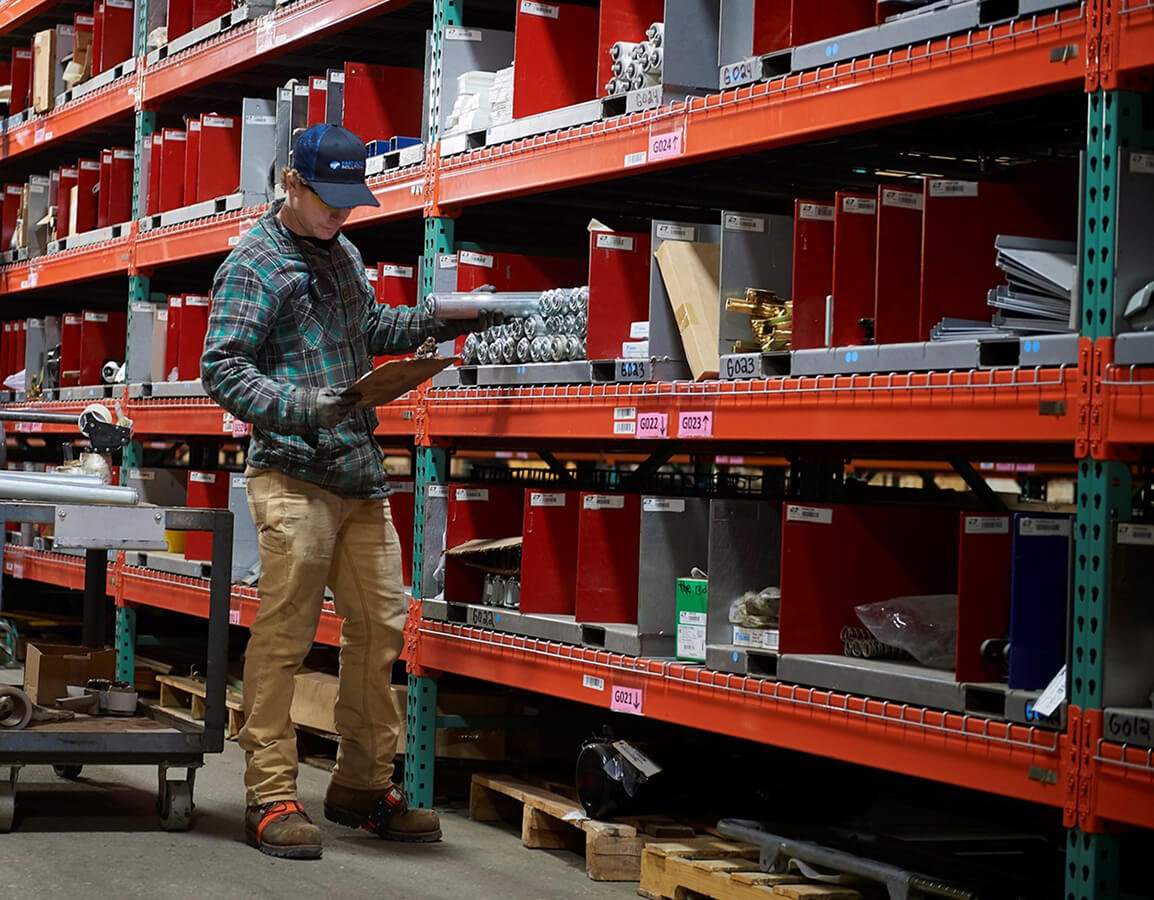 Warehouse worker with clipboard checking inventory on industrial shelving system filled with organized parts in red bins.