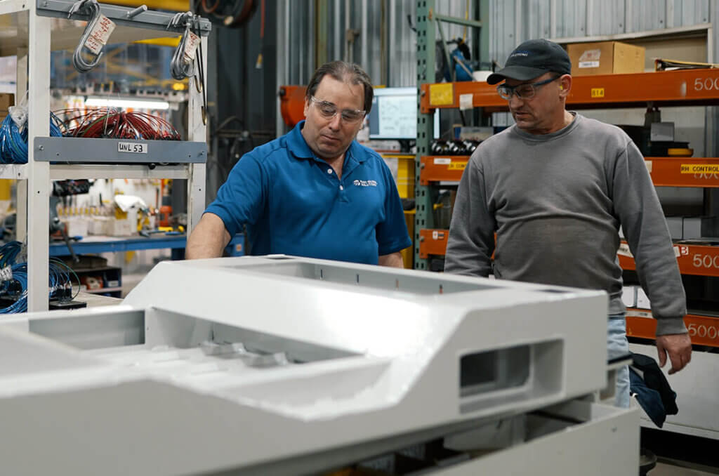 Two workers in safety glasses examining white metal enclosures in a factory with storage racks visible in the background.