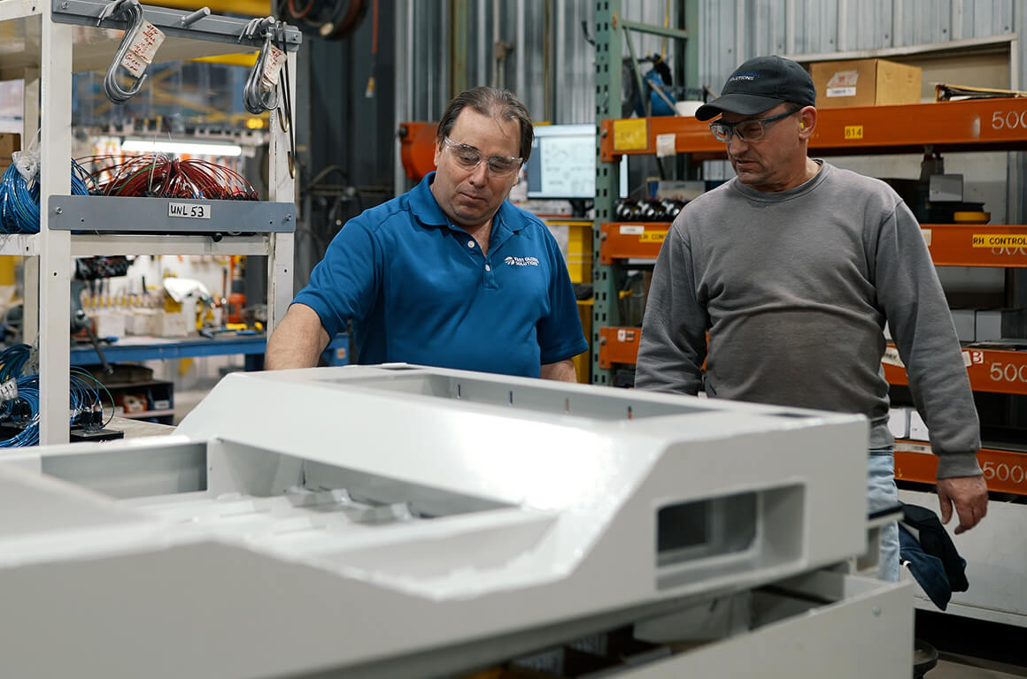 Two workers in safety glasses examining white metal enclosures in a factory with storage racks visible in the background.