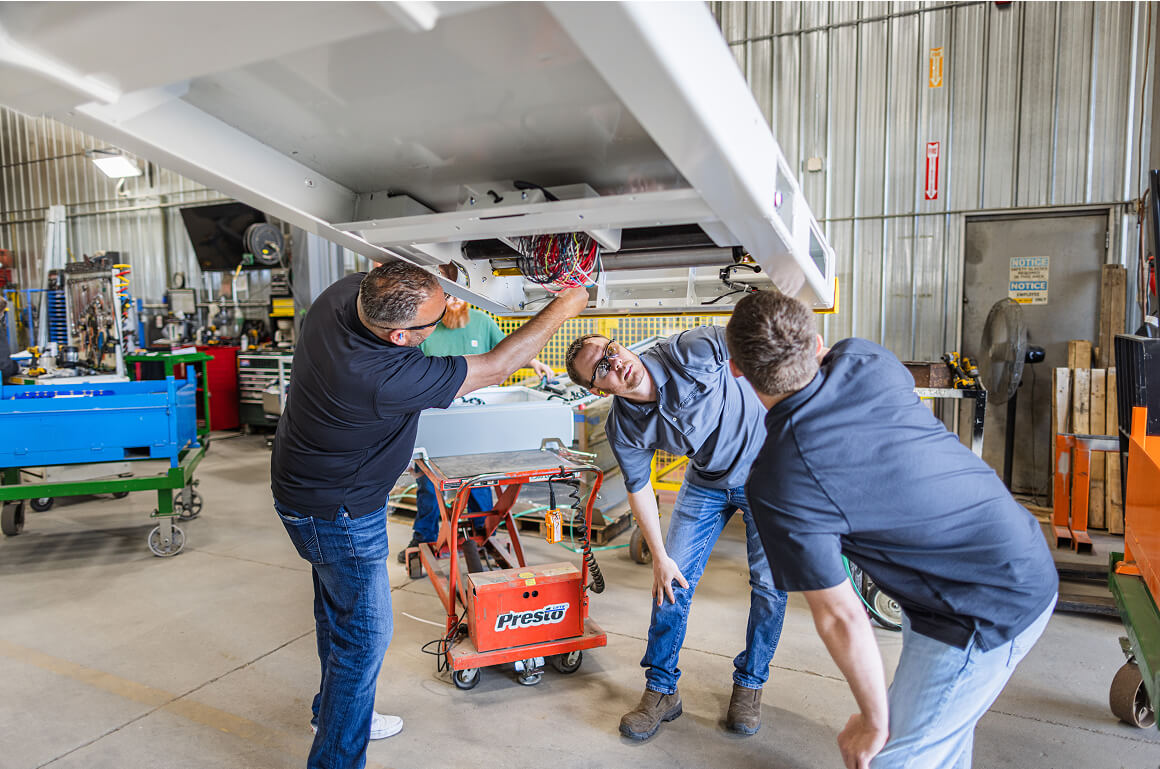 Three technicians working together to inspect and service the underside of an industrial conveyor system in a manufacturing facility.