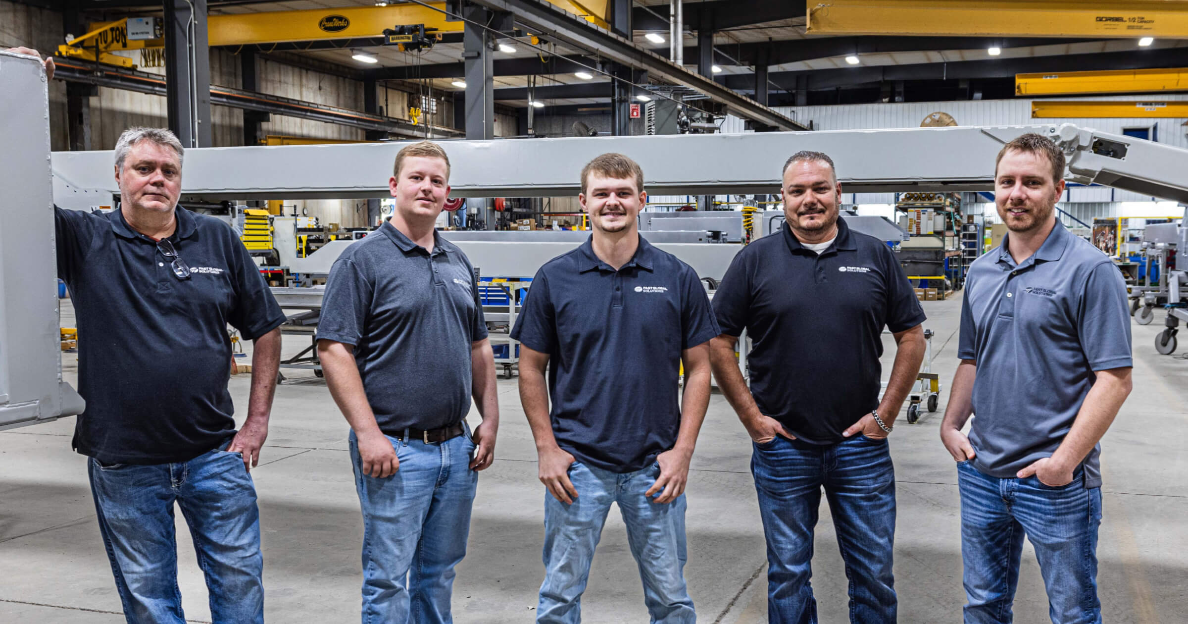 Five team members in navy polo shirts and jeans standing in manufacturing facility with conveyor equipment in background.