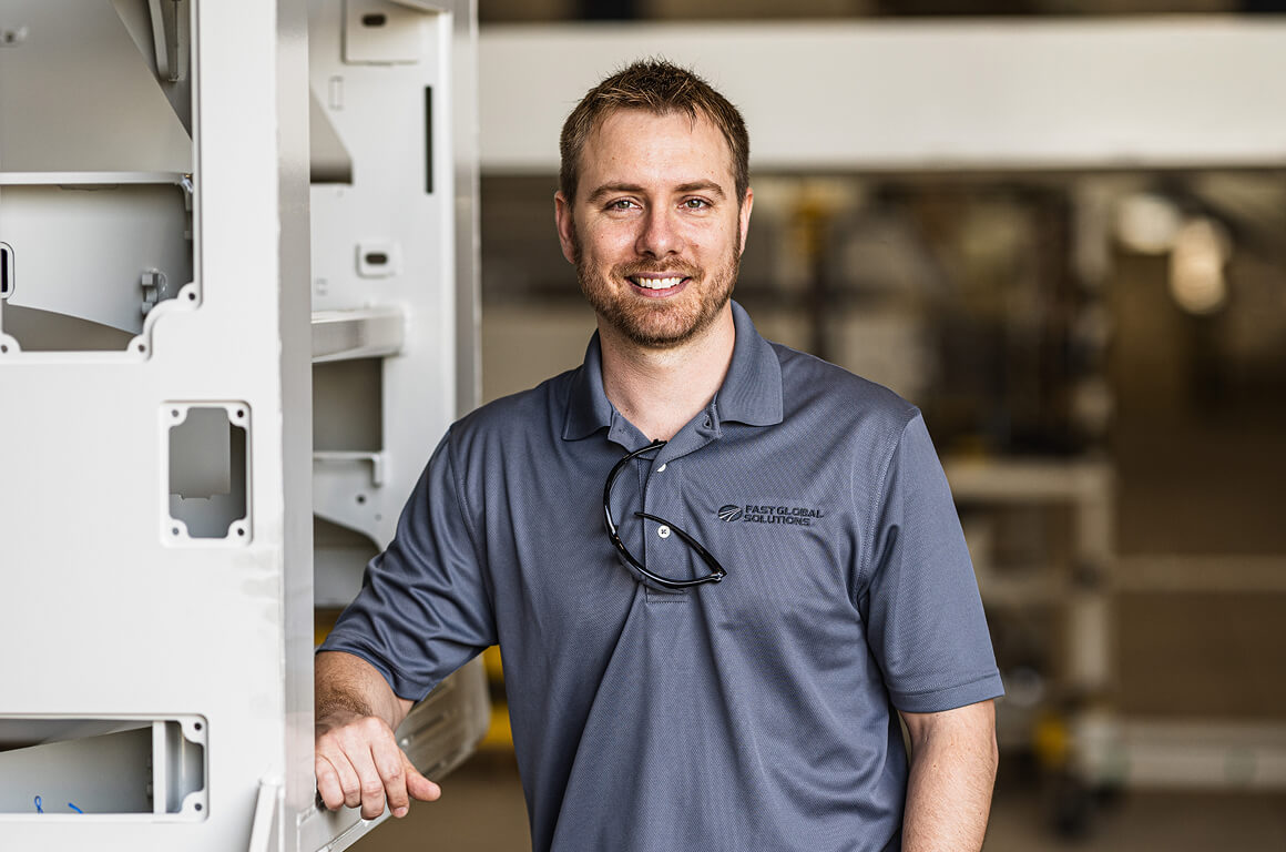 Smiling professional in a gray company polo shirt standing next to manufacturing equipment in a facility.