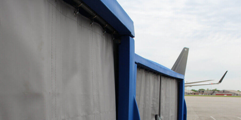Blue baggage cart with gray curtains on an airport tarmac with aircraft tail visible in background.