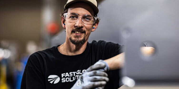 Person wearing safety equipment and FAST Global Solutions branded shirt working with manufacturing materials in a production environment.