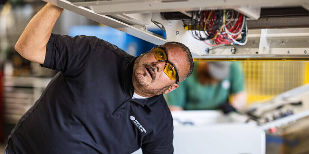 Worker in black polo shirt looking up at exposed wiring and electrical components during maintenance inspection of industrial conveyor equipment.