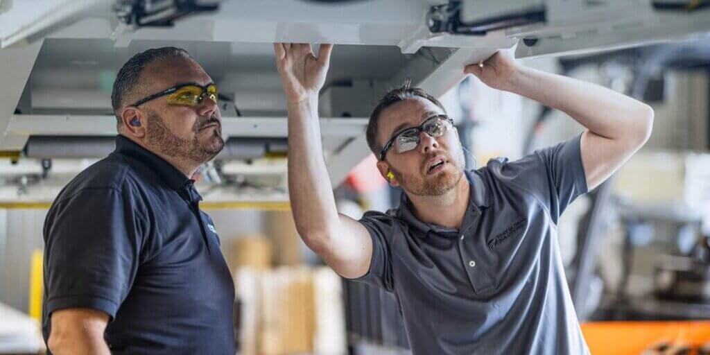 Two technicians in gray and navy polo shirts examining overhead equipment in manufacturing facility.